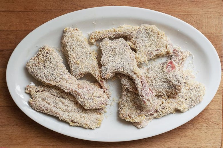A white plate of eight breaded lamb chops sits on a wooden surface.